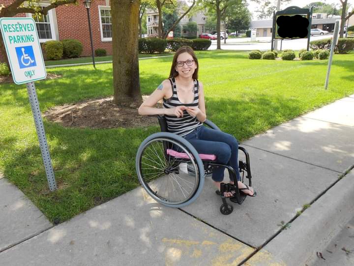 A white femme with brown hair and glasses sits in a wheelchair on a sidewalk, next to an accessible parking sign, and in front of a tree and brick building. They are wearing a black and grey tank top and jeans. 